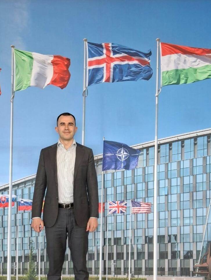 NAVO-medewerker Mike poseert voor een foto van een gebouw met wapperende vlaggen